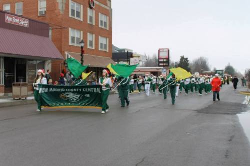 A parade featuring a marching band in green uniforms, holding banners and flags, walking down a snowy street.