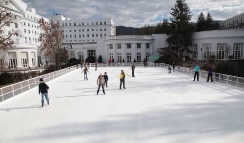 People ice skating on an outdoor rink surrounded by a large white building and trees under a cloudy sky.