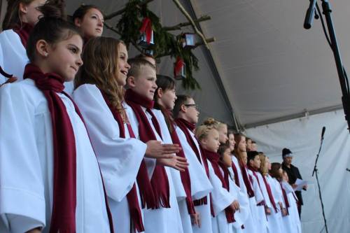 A children's choir in white robes and maroon scarves performs on stage under a tent, with a microphone in the foreground.