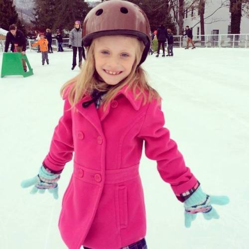 A smiling girl in a pink coat and helmet ice skating, with people skating in the background on a snowy rink.