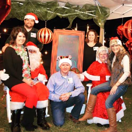 A festive group poses under a decorated tent, featuring Santa, holiday attire, and cheerful decorations.