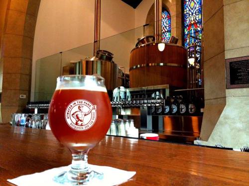 A glass of amber beer sits on a bar, with brewing equipment and stained glass windows in the background.