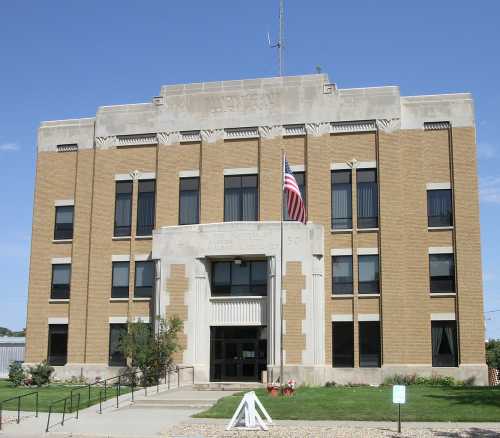 Historic brick building with a central entrance, flagpole, and decorative stonework, surrounded by greenery.