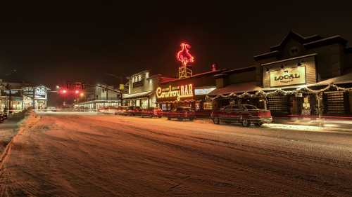 A snowy street at night featuring shops, a bar with a neon sign, and parked cars under festive lights.