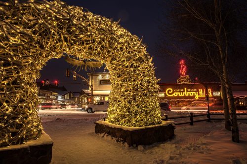 A festive arch made of lights and antlers, with a cowboy-themed sign in the background, set in a snowy night scene.