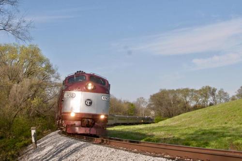 A vintage train approaches on a curved track, surrounded by green trees and a clear blue sky.