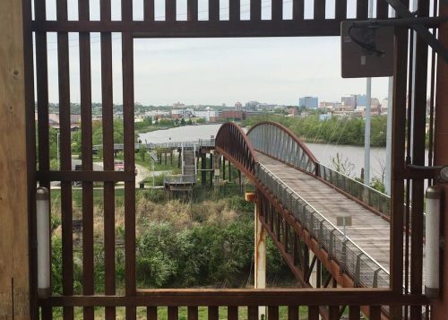 A view through a wooden frame of a bridge over a river, with city buildings in the background and greenery nearby.