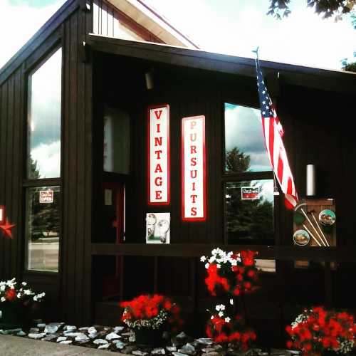 A vintage shop with a wooden exterior, red flowers, and an American flag displayed outside.