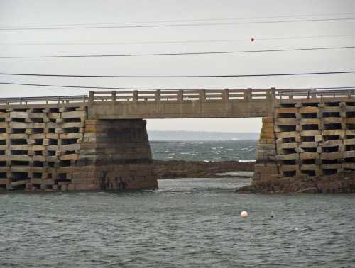 A concrete bridge spans over water, supported by stone pillars, with a cloudy sky in the background.