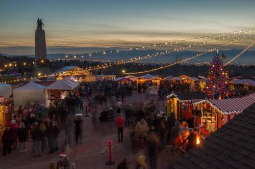 A festive market scene at dusk, with twinkling lights, a Christmas tree, and crowds enjoying the holiday atmosphere.