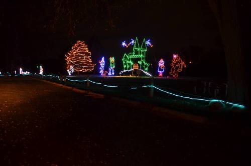 Colorful holiday lights illuminate a park scene, featuring a castle, trees, and festive characters against a dark sky.