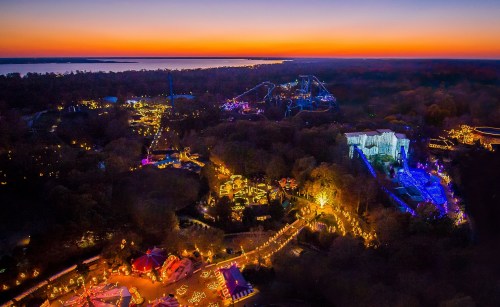 Aerial view of a vibrant amusement park at sunset, illuminated with colorful lights against a darkening sky.