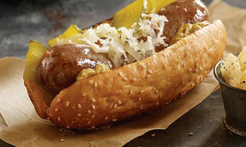 A sesame seed bun filled with two sausages, topped with mustard, sauerkraut, and pickles.