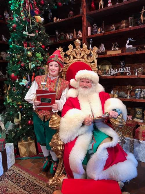 A cheerful Santa Claus sits on a golden throne beside an elf holding a gift, with a decorated Christmas tree in the background.