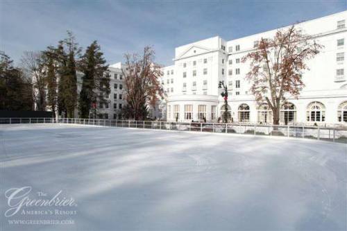 A snowy ice rink in front of a large white hotel surrounded by trees.