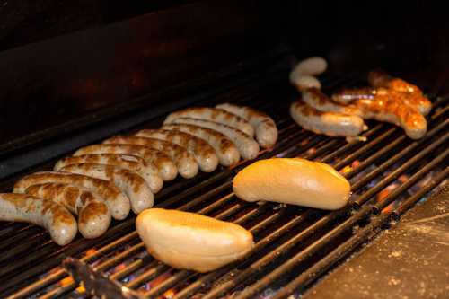 Grilled sausages and buns cooking on a barbecue grill, with flames visible beneath.