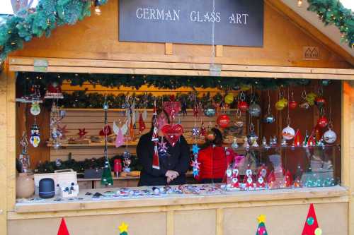 A festive market stall displaying colorful German glass art ornaments, with two people behind the counter.