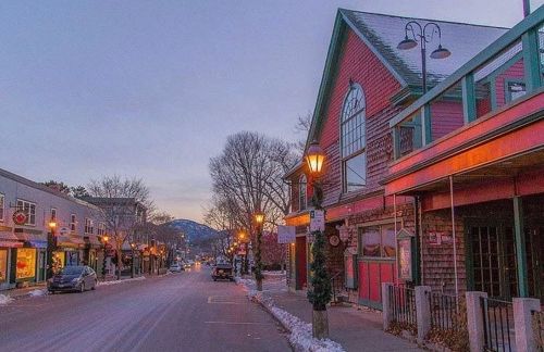 A quiet, snow-covered street at dusk, lined with charming shops and warm streetlights, with mountains in the background.