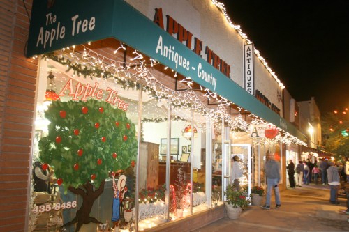 A festive storefront of "The Apple Tree" antique shop, decorated with lights and a tree mural, bustling with evening shoppers.