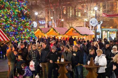 A festive crowd gathers around holiday stalls and a brightly lit Christmas tree in a winter market.