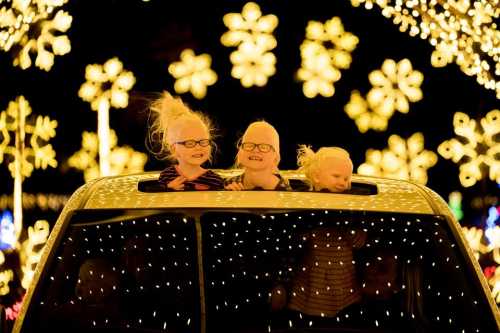 Three children with glasses smile from the sunroof of a car, surrounded by bright, festive holiday lights.