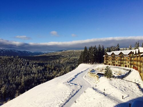 A snowy landscape with a mountain resort nestled among evergreen trees under a clear blue sky.
