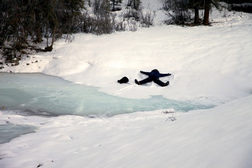 A person in winter clothing lies in the snow near a frozen stream, surrounded by trees and a snowy landscape.