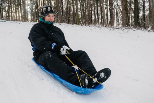 A person in winter clothing sleds down a snowy hill, smiling while holding onto the sled's rope.
