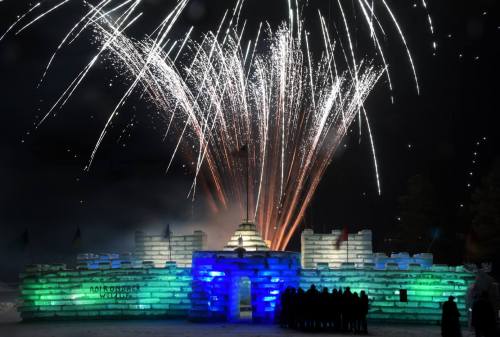 A colorful ice castle illuminated by fireworks, with a crowd silhouetted in front, celebrating at night.