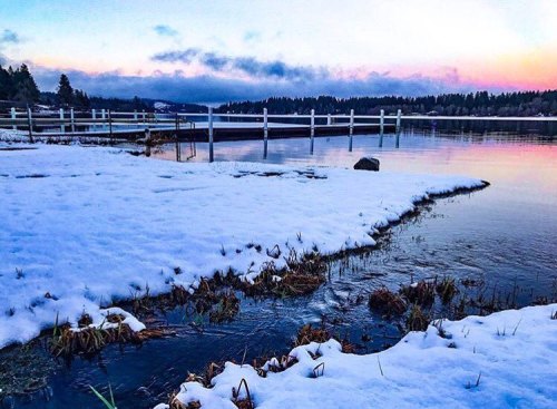A serene winter landscape with snow-covered ground, a calm lake, and a colorful sunset in the background.