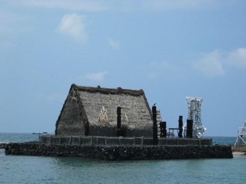A thatched-roof structure on a rocky shore, surrounded by water, with tall statues nearby under a blue sky.