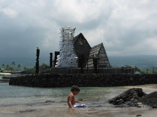 A young boy wades in shallow water near a beach with traditional huts and a cloudy sky in the background.