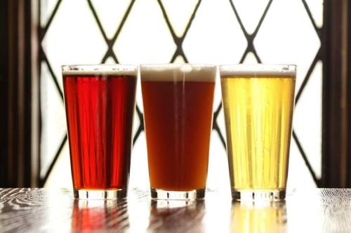 Three glasses of beer in varying colors: red, amber, and golden, set against a window with a geometric pattern.
