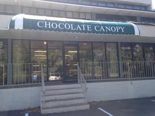 Exterior of a shop called "Chocolate Canopy," featuring a green awning and large windows, with a sign indicating it's open.