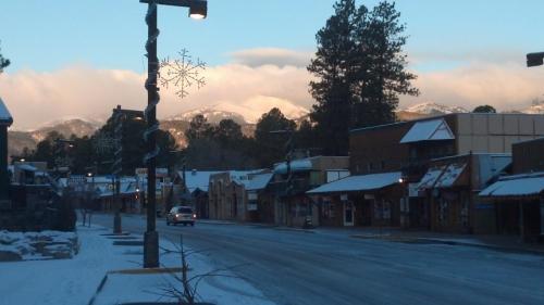 Snow-covered street with shops, mountains in the background, and a clear blue sky. Snowflake decoration on a lamp post.