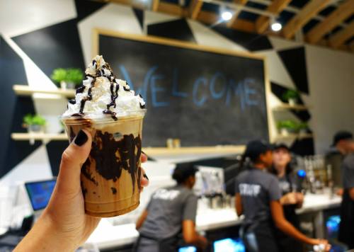 A hand holds a chocolate dessert drink topped with whipped cream and chocolate drizzle, with a café backdrop.