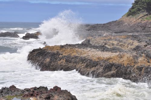 Waves crash against rocky coastline under a cloudy sky, creating a misty spray over the shoreline.