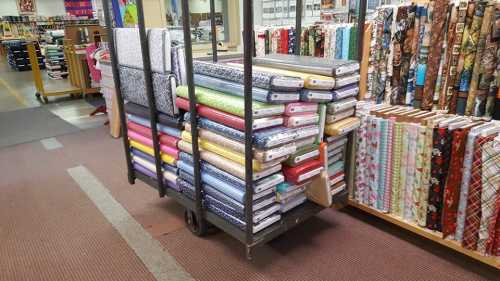 A cart filled with colorful fabric rolls in a craft store, surrounded by shelves of more fabric.