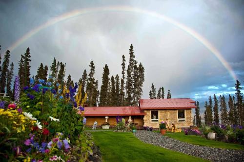 A colorful garden with flowers in front of a rustic house, under a vibrant rainbow and surrounded by tall trees.