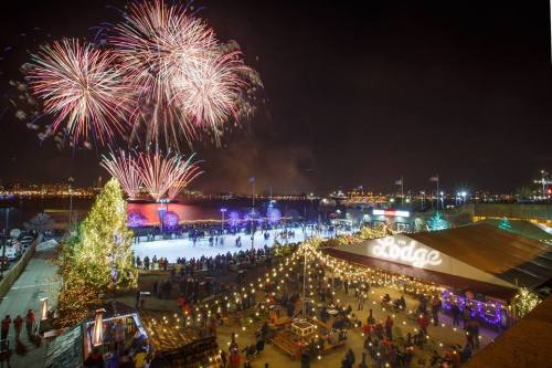 Fireworks illuminate the night sky over a festive ice skating rink and a cozy lodge, surrounded by holiday lights.
