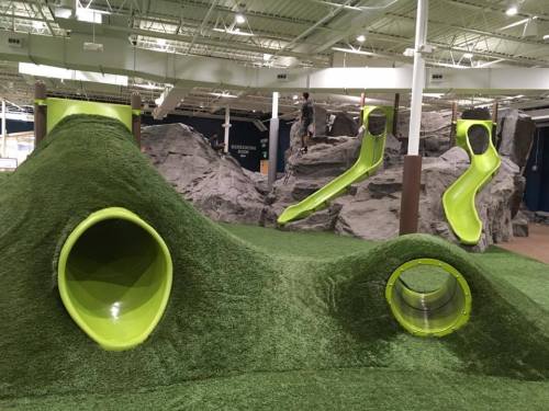 Indoor playground featuring green slides and climbing structures resembling hills and rocks.