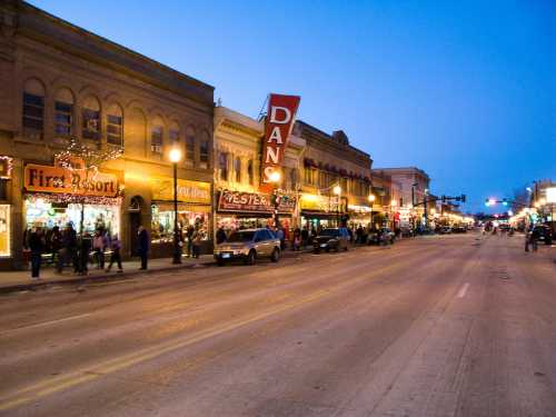 A lively street scene at dusk, featuring shops and restaurants with bright lights and a clear sky.