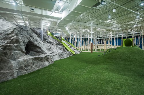 Indoor play area featuring artificial grass, slides, rock formations, and wooden structures for children to explore.