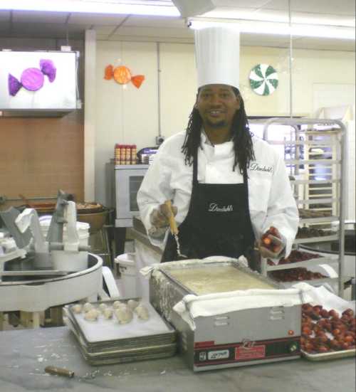 A chef in a white uniform and hat prepares food in a kitchen, surrounded by baking trays and colorful candy decorations.