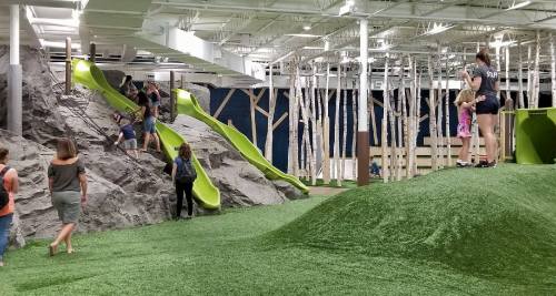 Indoor play area featuring green slides, artificial grass, and children climbing on rock-like structures.