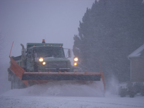 A snowplow clears a road during a heavy snowstorm, with visibility reduced and snow swirling around.