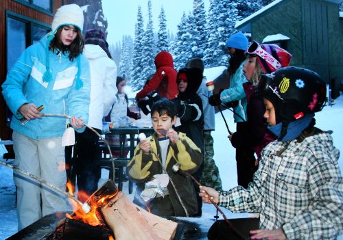 Children roasting marshmallows over a fire in a snowy outdoor setting, wearing winter clothing and enjoying the moment.