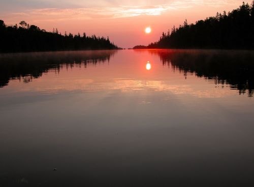 A serene lake at sunset, reflecting vibrant colors and surrounded by trees, with mist rising from the water's surface.