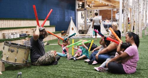 A group of children and adults play with colorful sticks in a playful music session in a bright, indoor space.