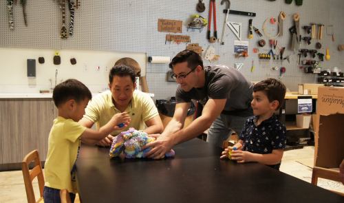 Two adults and two children gather around a table, playing with colorful toys in a workshop setting.